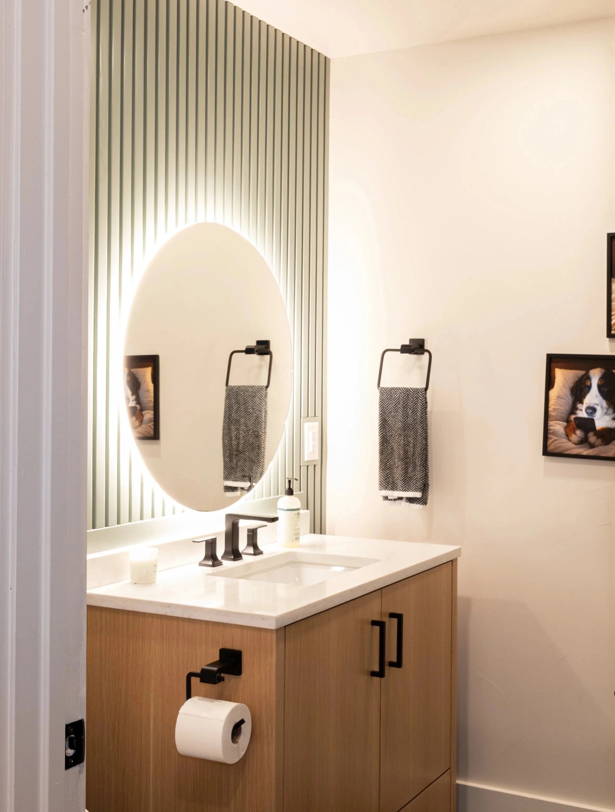 Modern bathroom with a wooden vanity, white countertop, oval mirror with LED lighting, black faucet, two black wall-mounted towel holders with towels, framed photo of a dog on the wall, and a toilet paper roll.