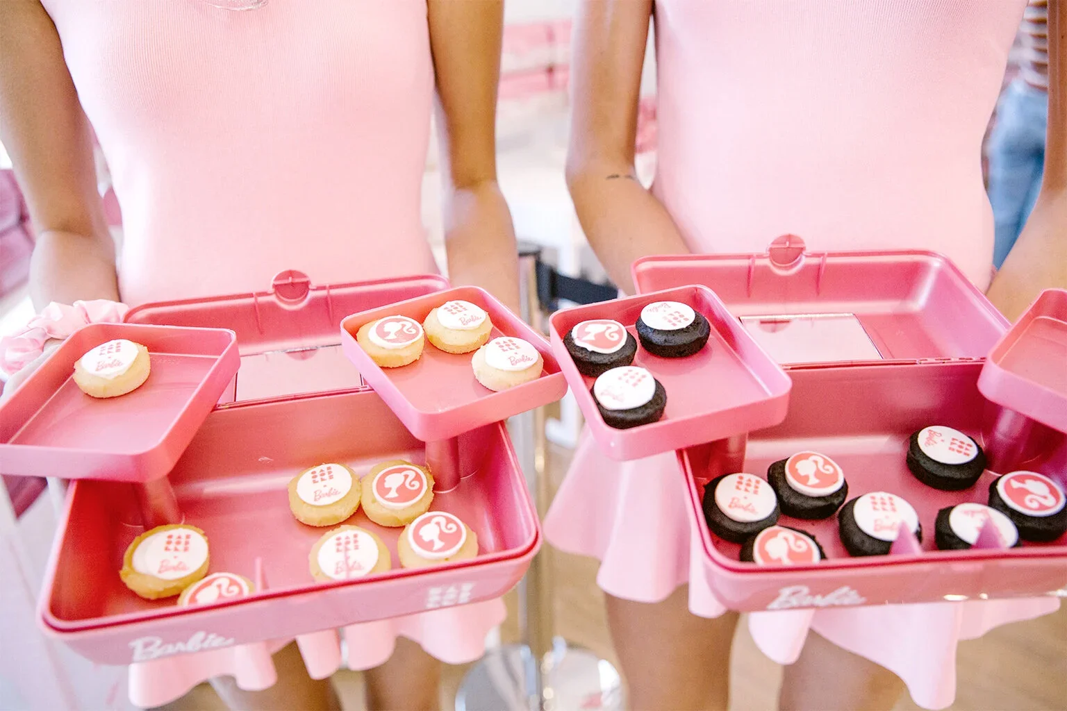 Two people in pink dresses holding pink trays with Barbie-themed cookies decorated with pink, white, and black icing and Barbie logos.
