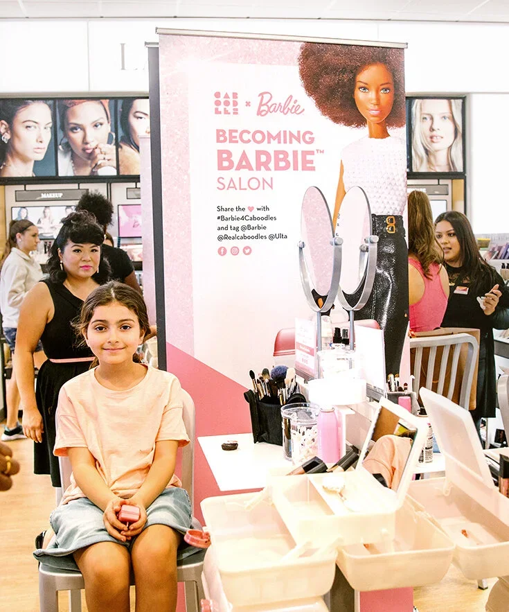 A young girl sitting in a makeup chair at the Barbie Becoming Barbie Salon, surrounded by makeup and beauty products, with a large Barbie advertisement behind her and other people in the background.