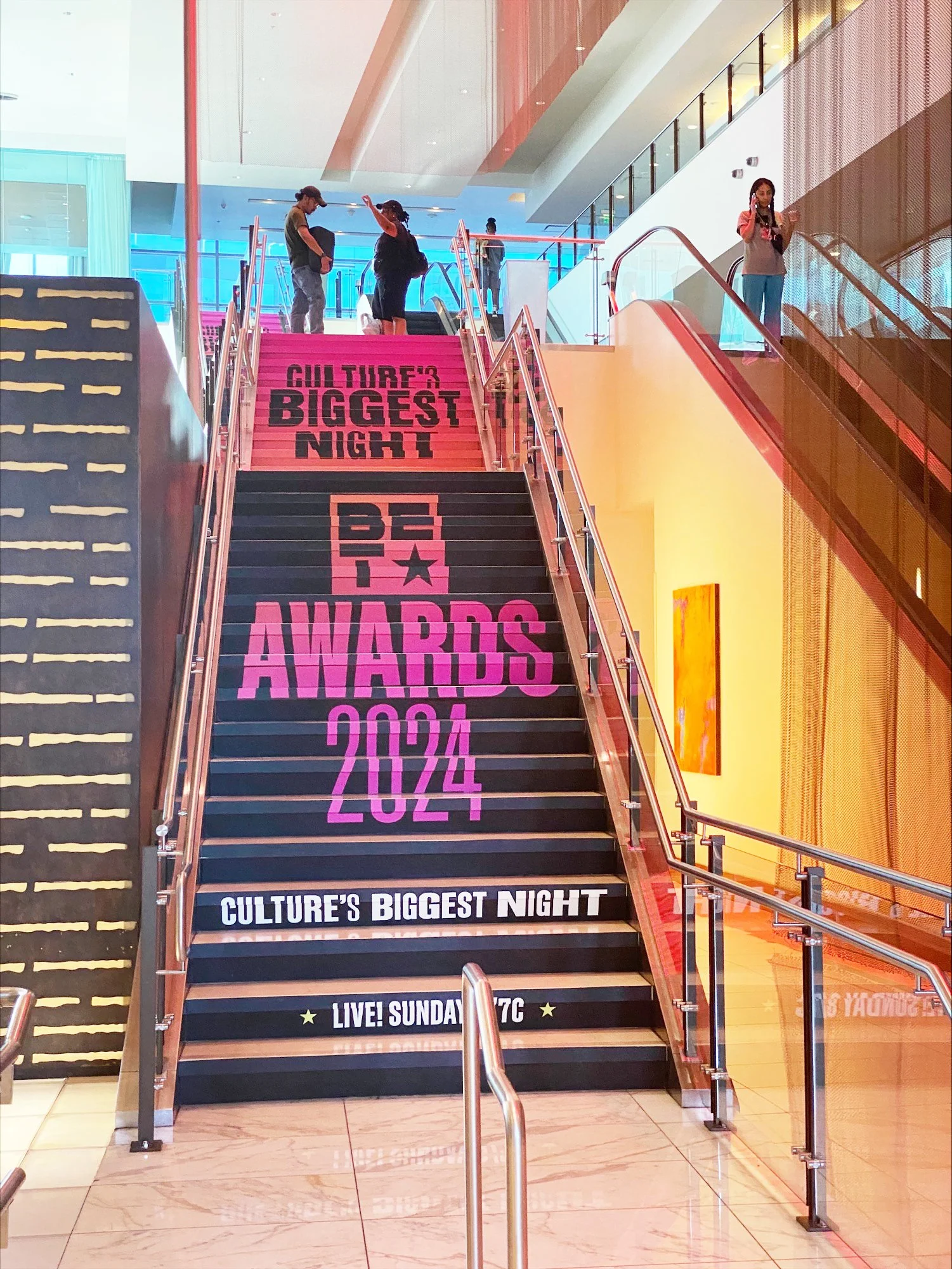 Staircase in a shopping mall decorated with colorful banners advertising the 2024 Culture's Biggest Night Awards event, with people ascending and descending the stairs.