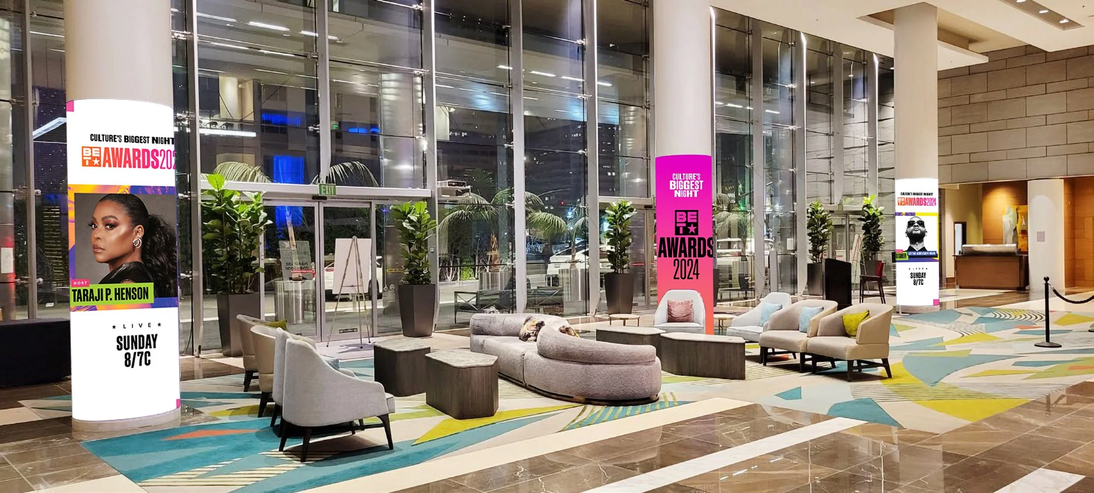 Lobby or reception area of an event venue with digital banners advertising the Be The Awards 2024 event, featuring a woman named Taraji P. Henson and a man with a beard and sunglasses, arranged with chairs and plants near large glass windows.
