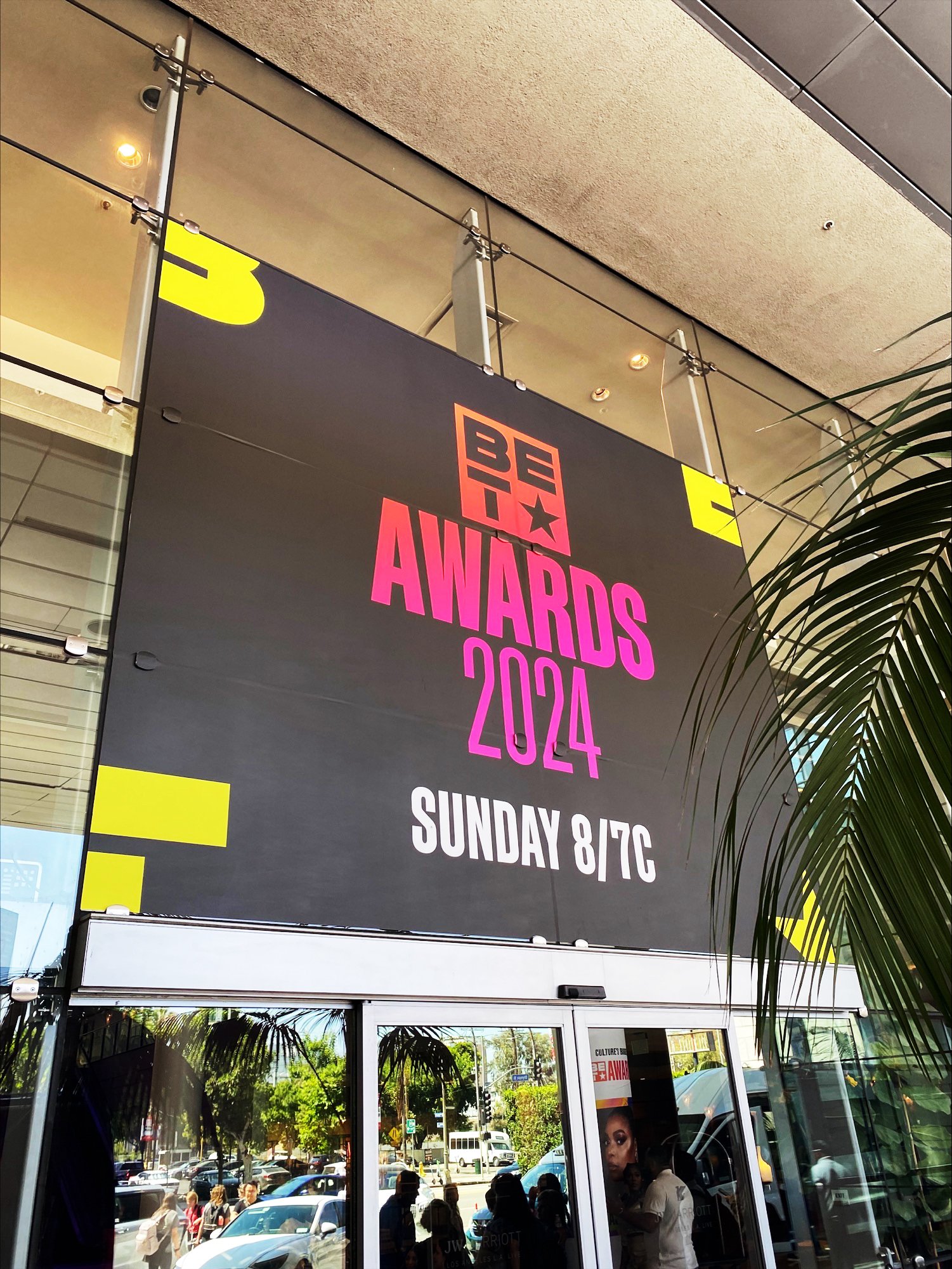 Banner announcing the 2024 Awards show on Sunday, August 4th at 8/7c, displayed at a building entrance. The reflection in the glass shows a busy street with cars and pedestrians.