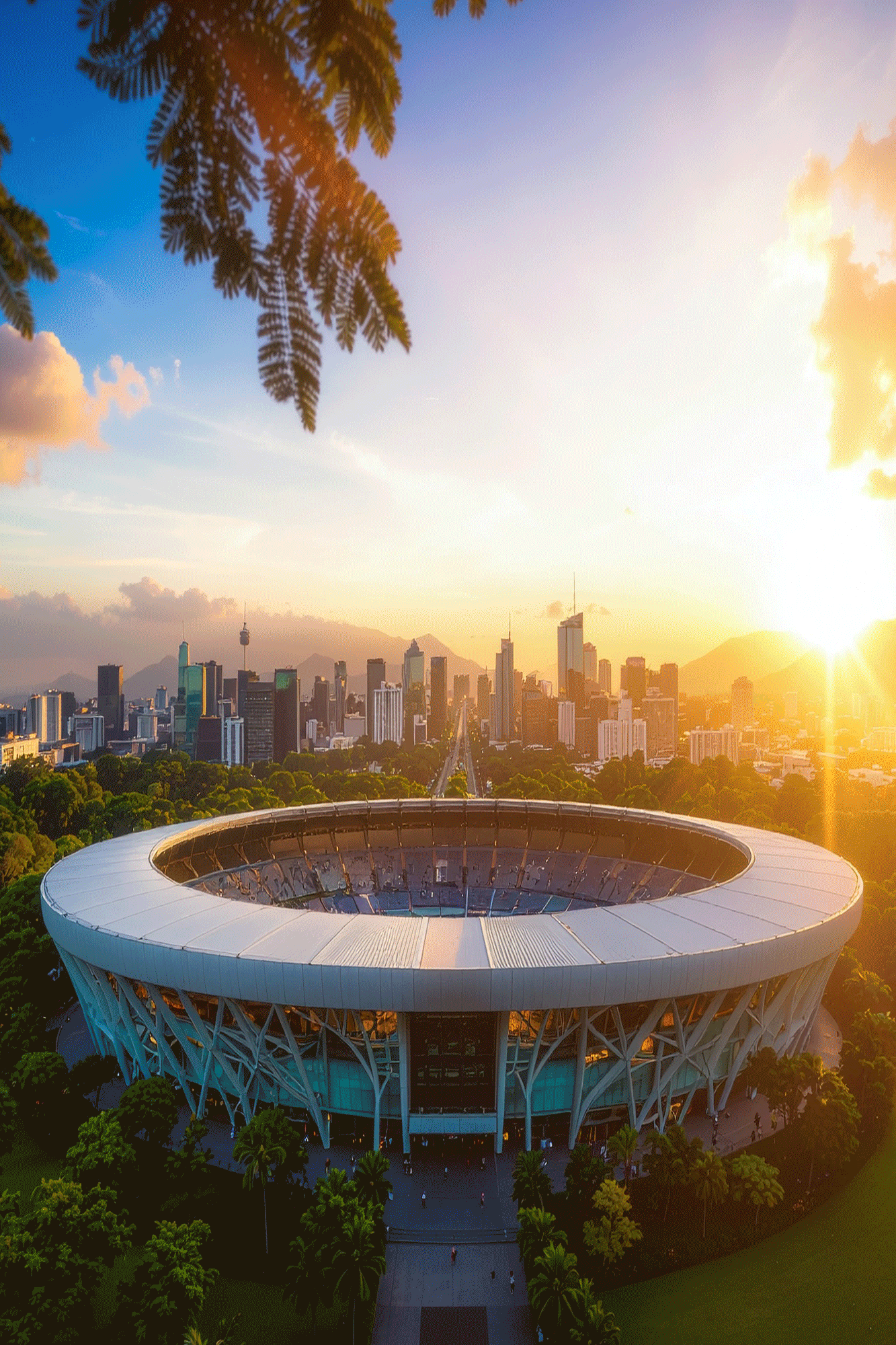 A city skyline at sunset viewed from a tall tower with a circular observation deck in the foreground. Trees surround the tower, and tall buildings stretch across the horizon with mountains in the background. The sky is partly cloudy with the sun setting on the right.