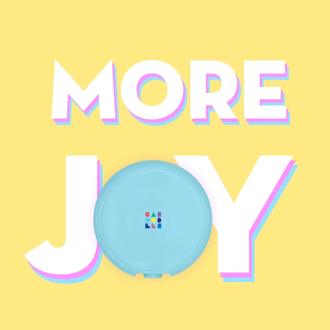 A yellow background with the words 'MORE JOY' in large white letters with a pink and blue shadow effect. A light blue circular object with colorful small blocks in the center is placed over the letter 'O' in 'JOY'.