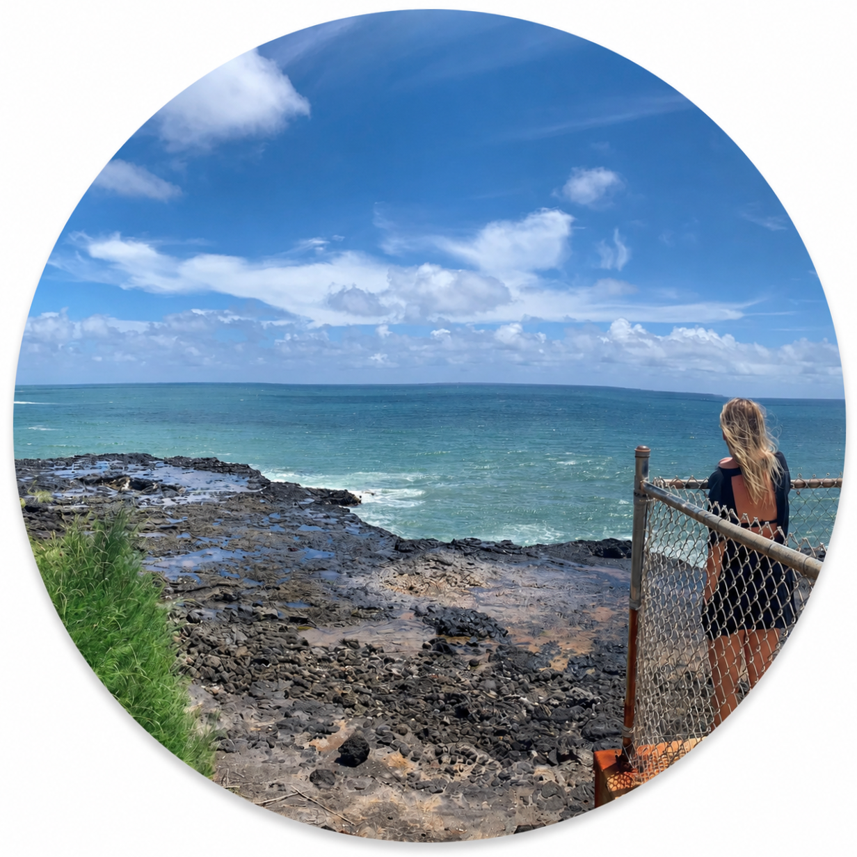 A woman with long blonde hair looking out over the ocean from a rocky shoreline behind a chain-link fence on a sunny day with blue sky and scattered clouds.