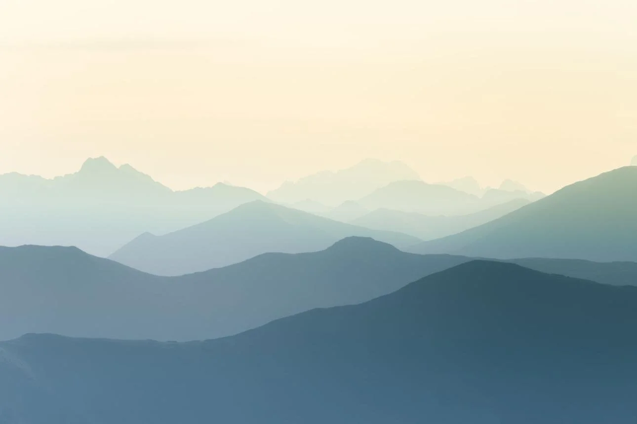 Silhouettes of mountain ranges fading into the distance with a pale sky in the background.