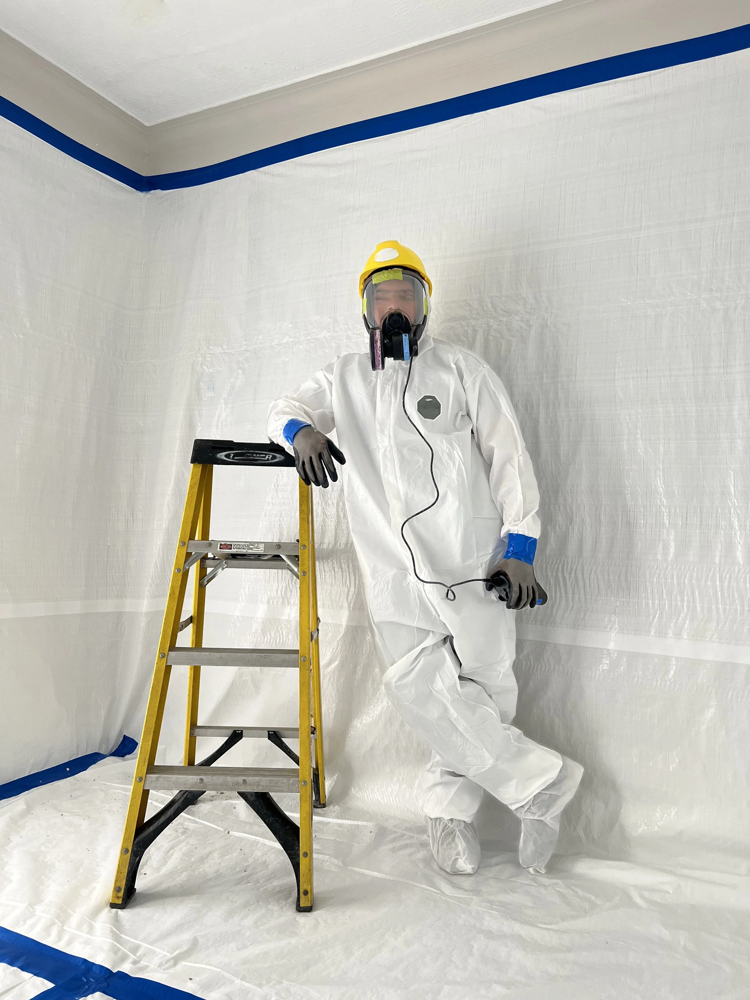 A person in protective gear, including a yellow hard hat, face mask, gloves, and a full-body white suit, standing next to a yellow ladder and holding a tool, inside a room with white walls and plastic sheeting covering the floor and walls for painting or renovation.