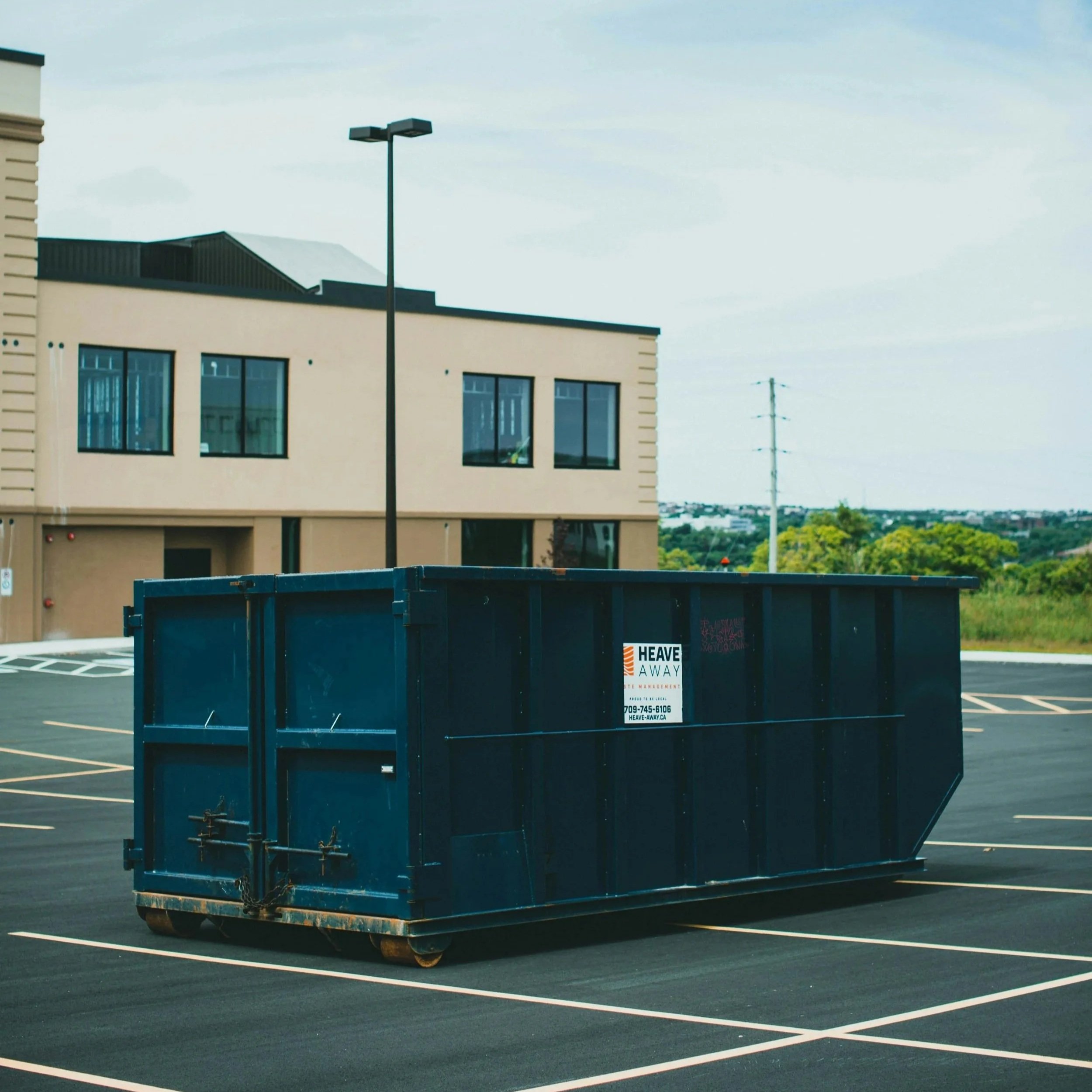 A large blue dumpster with a sign that says 'Heave Away' in a parking lot. In the background are a beige building with large windows and a streetlight. The sky is partly cloudy.