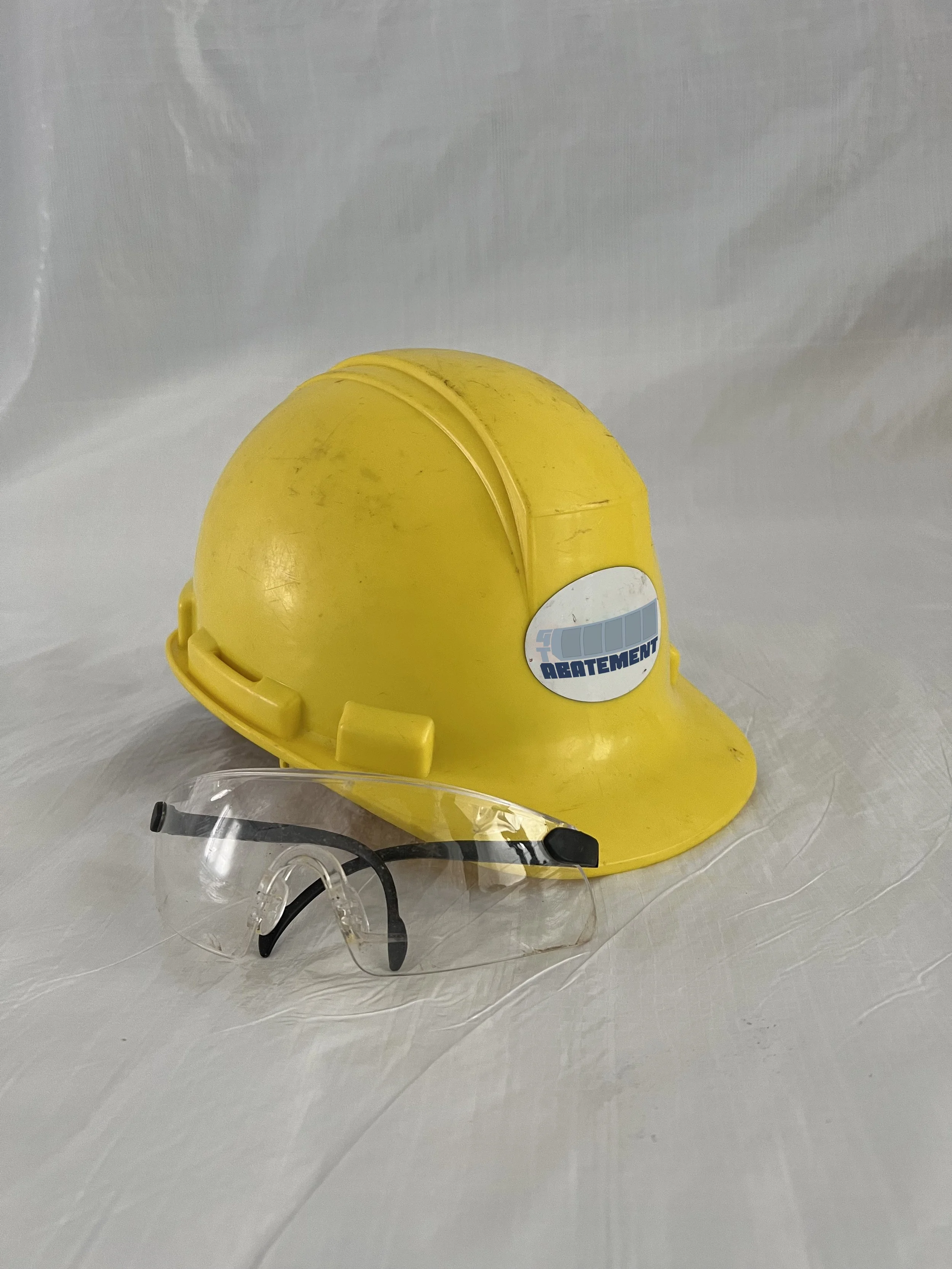 A yellow construction helmet with a sticker that reads 'Top Drafting' and a clear safety face shield placed in front of it on a white surface.