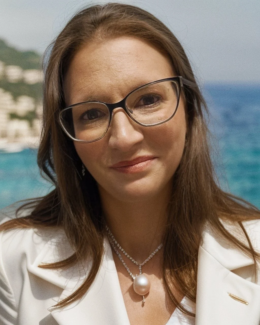 A woman with brown hair and glasses smiling outdoors with a body of water and buildings in the background.