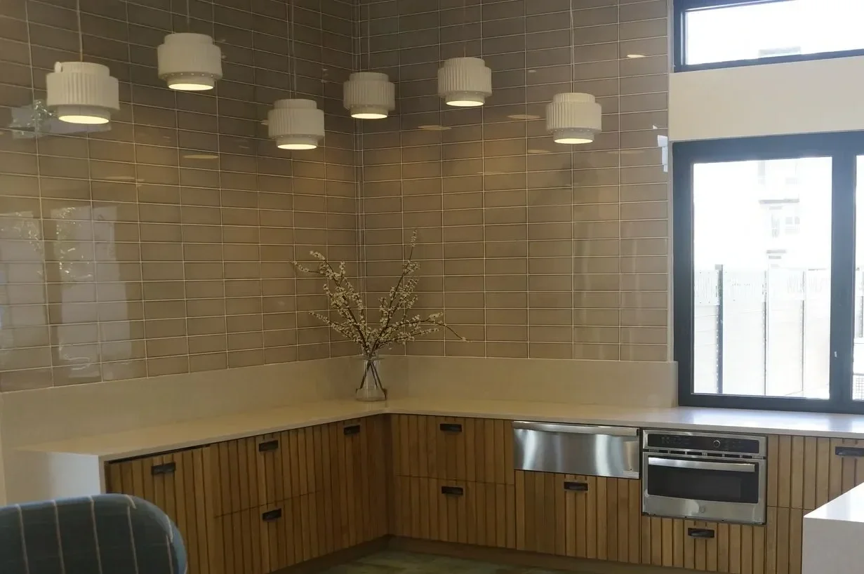 Modern kitchen with beige tiled walls, wooden cabinetry, and a window with black trim. Decorative branches in a vase on the countertop, which also features a built-in oven and dishwasher, illuminated by white pendant lights.