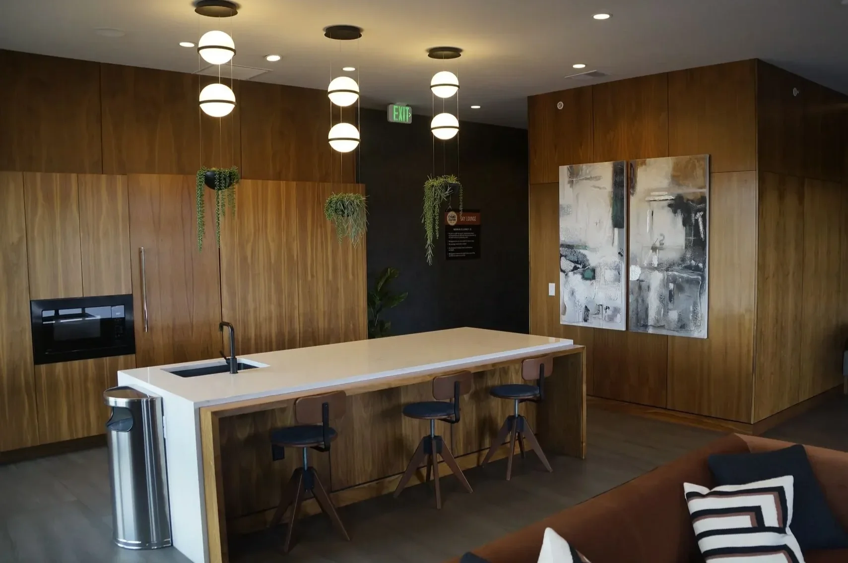 Modern kitchen with wooden cabinets, a white island with a built-in microwave and sink, hanging pendant lights, decorative plants, and abstract art on the wall.