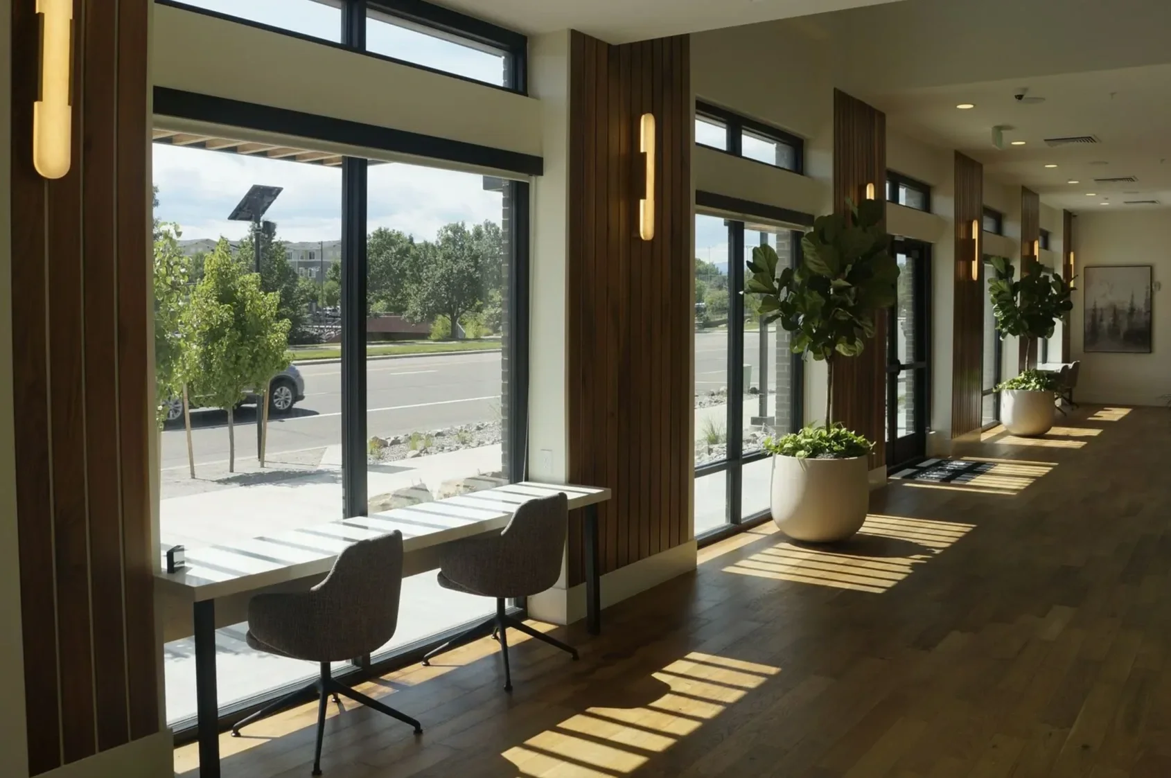 Modern interior with large windows, potted plants, and wooden wall panels, sunlight casting shadows on the wooden floor.