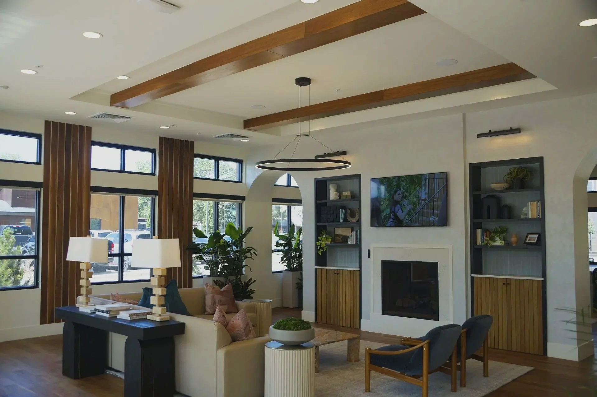 Modern living room with large windows, wooden beams on the ceiling, a white fireplace, built-in shelves with decor, a flat-screen TV, a beige sofa with colorful pillows, a black side table with books and candle holders, and indoor plants.
