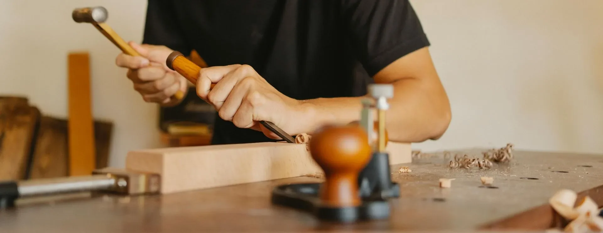 A person woodworking, carving a piece of wood with a chisel and hammer, surrounded by woodworking tools and wood shavings on a worktable.