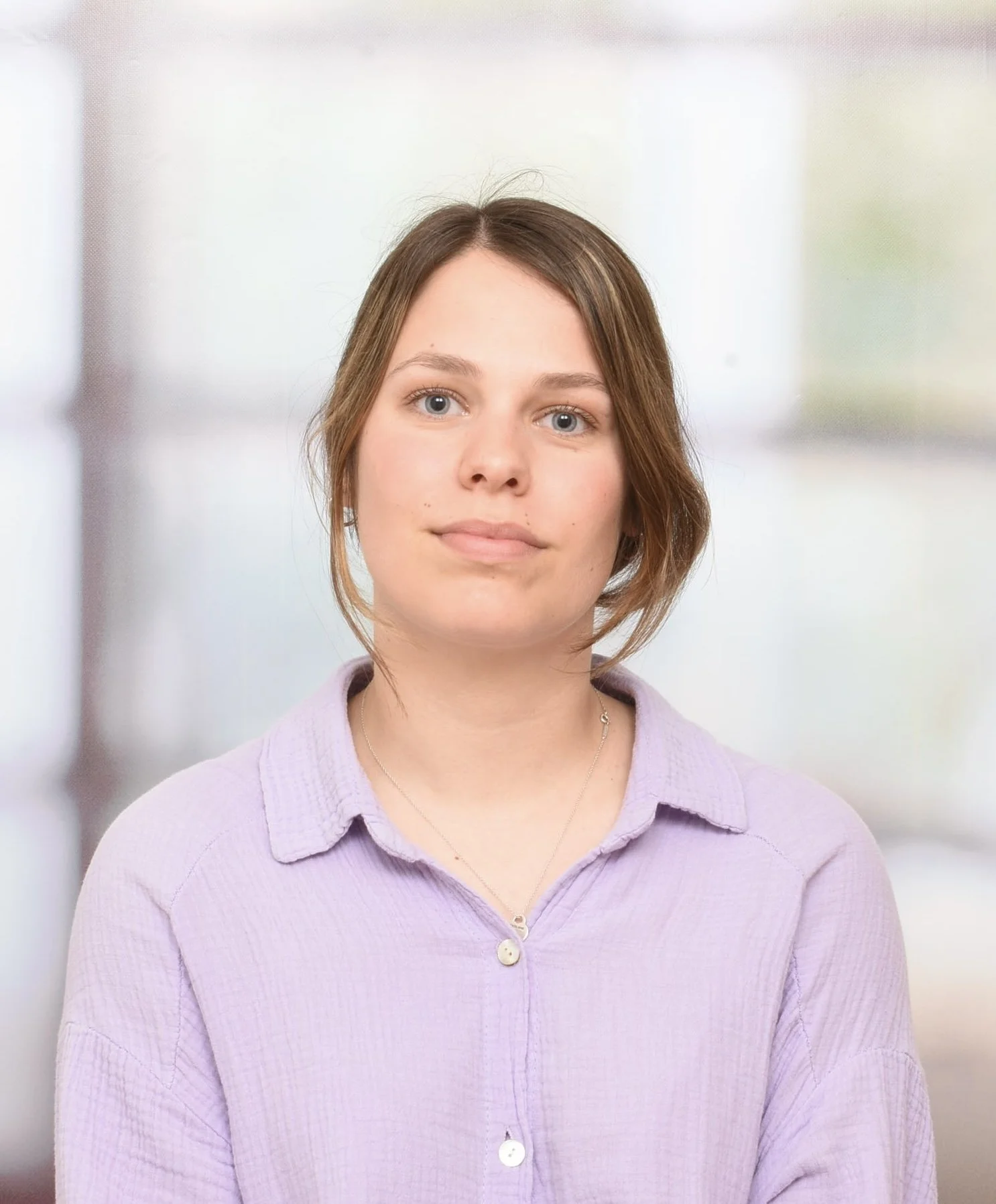 A young woman with short brown hair wearing a light purple button-up shirt and necklace, standing in a brightly lit indoor space with a blurred background.