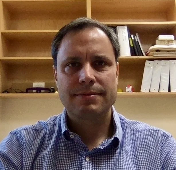 A man with short dark hair wearing a blue checked shirt taking a selfie in front of a wooden bookshelf with files and books.