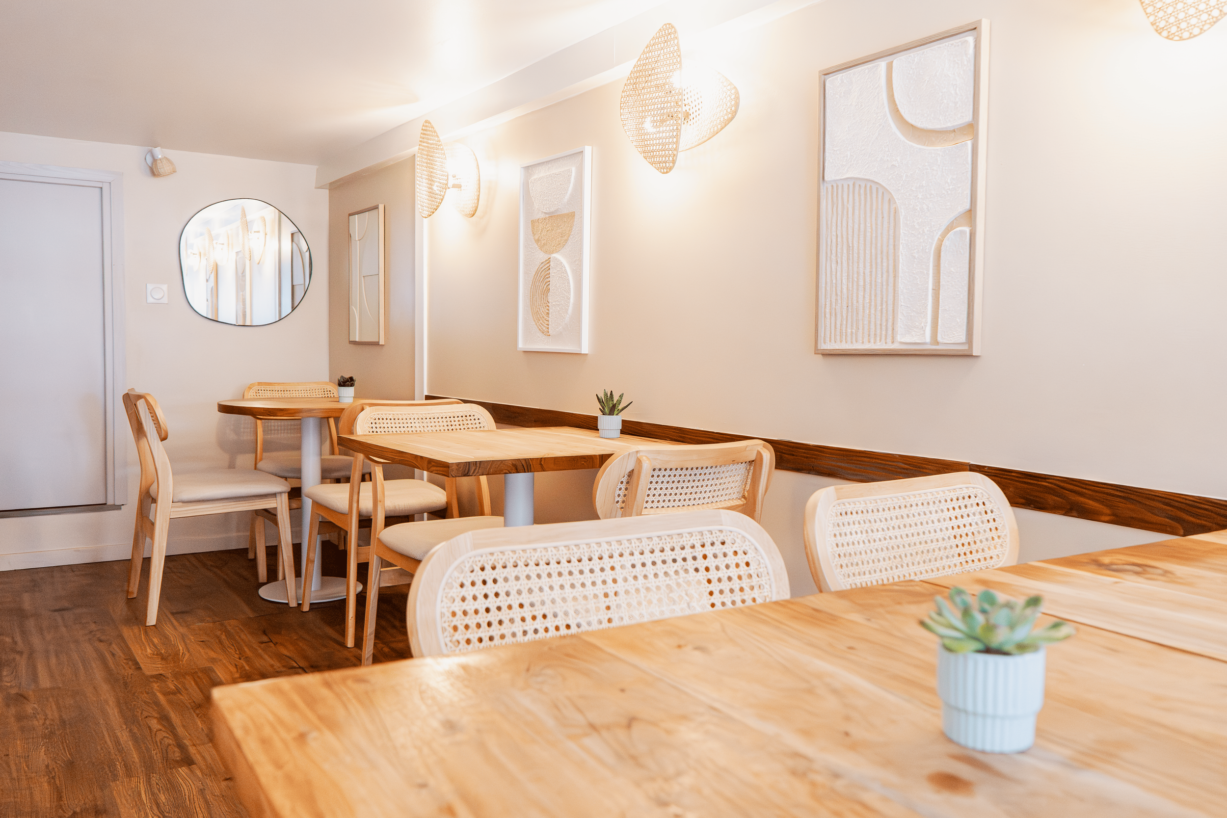 Intérieur d'une salle à manger de chez Xocolatte à Lille aux murs beige, meules en bois clair, avec des tableaux abstraits, un miroir rond et des lampes en rotin.