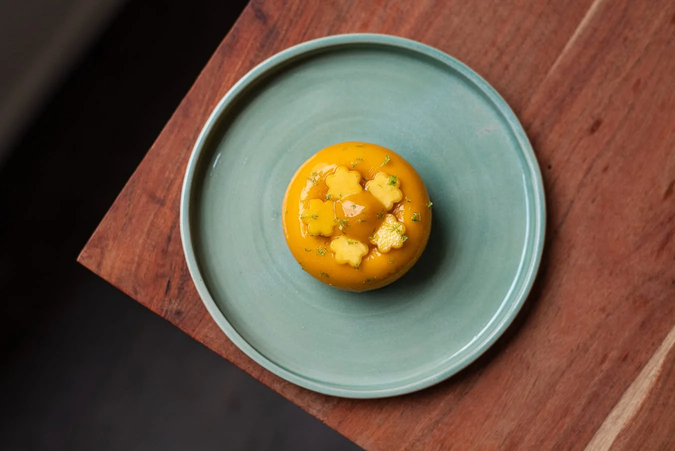 Dessert jaune avec des fleurs en pâte décoratives et un sirop, présenté dans une assiette bleue sur une table en bois de chez Xocolatte à Lille