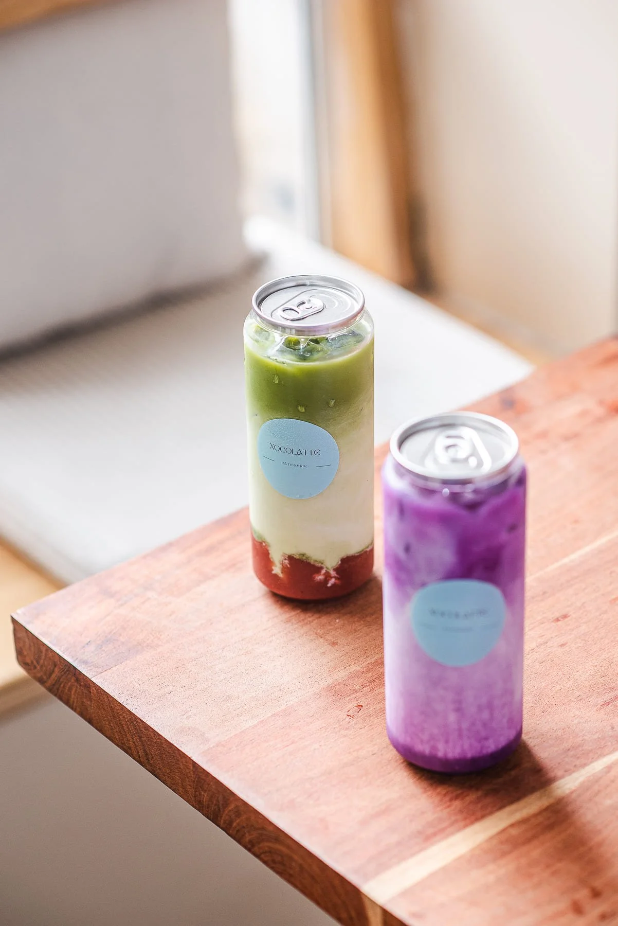 Deux canettes de boissons colorées, de chez Xocolatte à Lille ,une verte et une violette, posées sur une table en bois avec un fond flou.