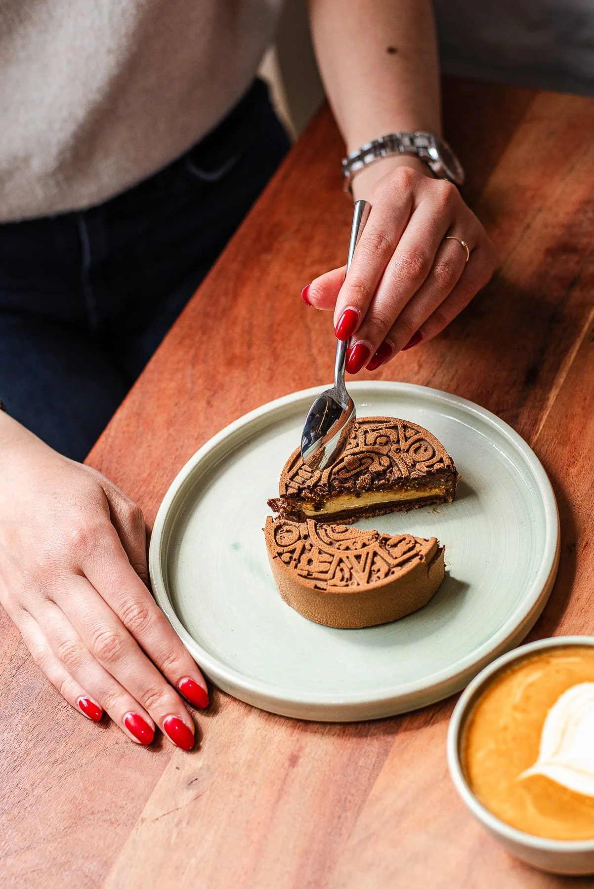 Une personne avec des ongles rouges mange un gâteau au chocolat de chez Xocolatte à Lille. La personne porte une montre et une alliance dorée, et la photo est prise sur une table en bois.