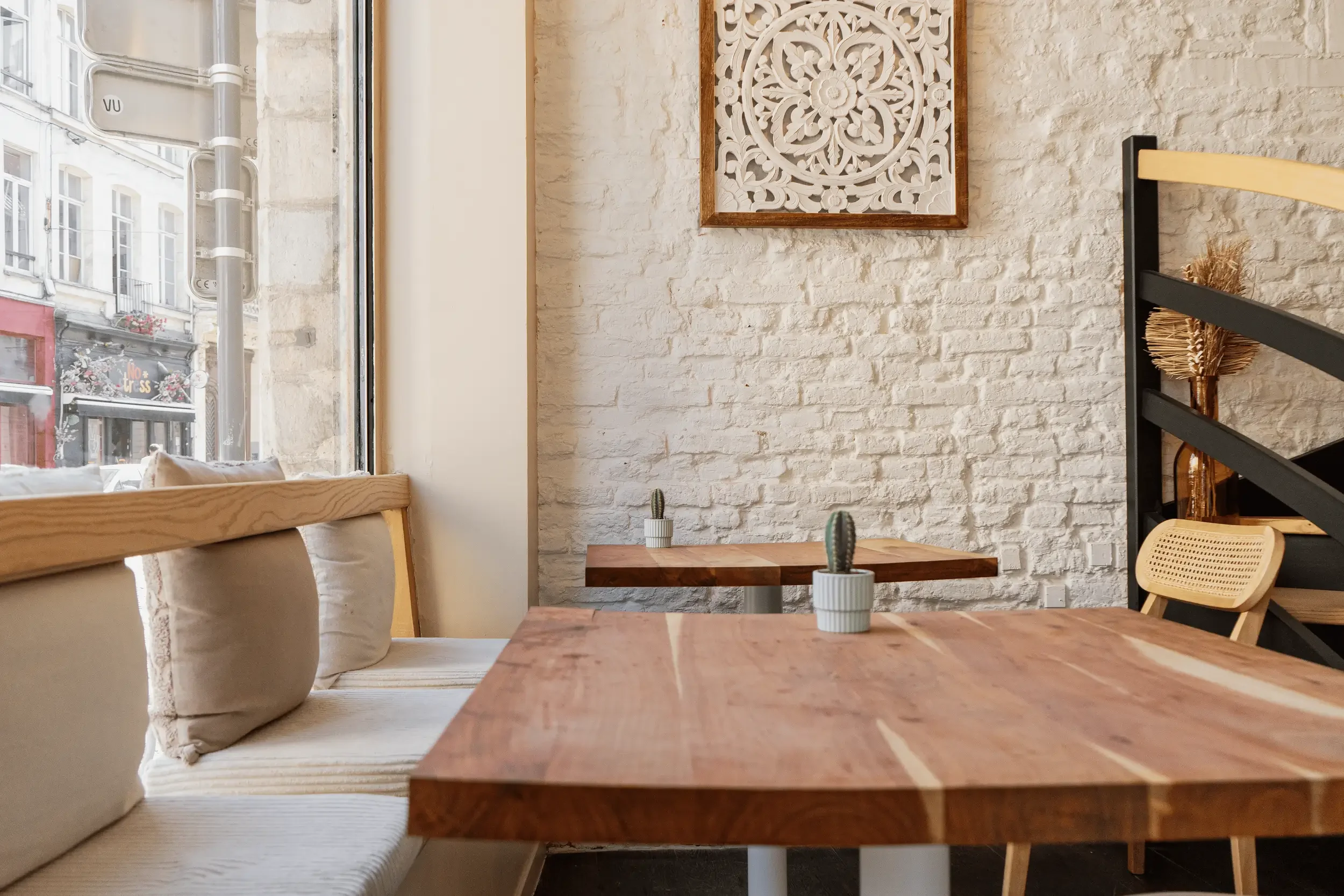 Intérieur de chez Xocolatte à Lille, avec tables en bois, coussins beige, et murs en pierre blanche. Une grande fenêtre laisse entrer la lumière naturelle et offre une vue sur la rue extérieure.
