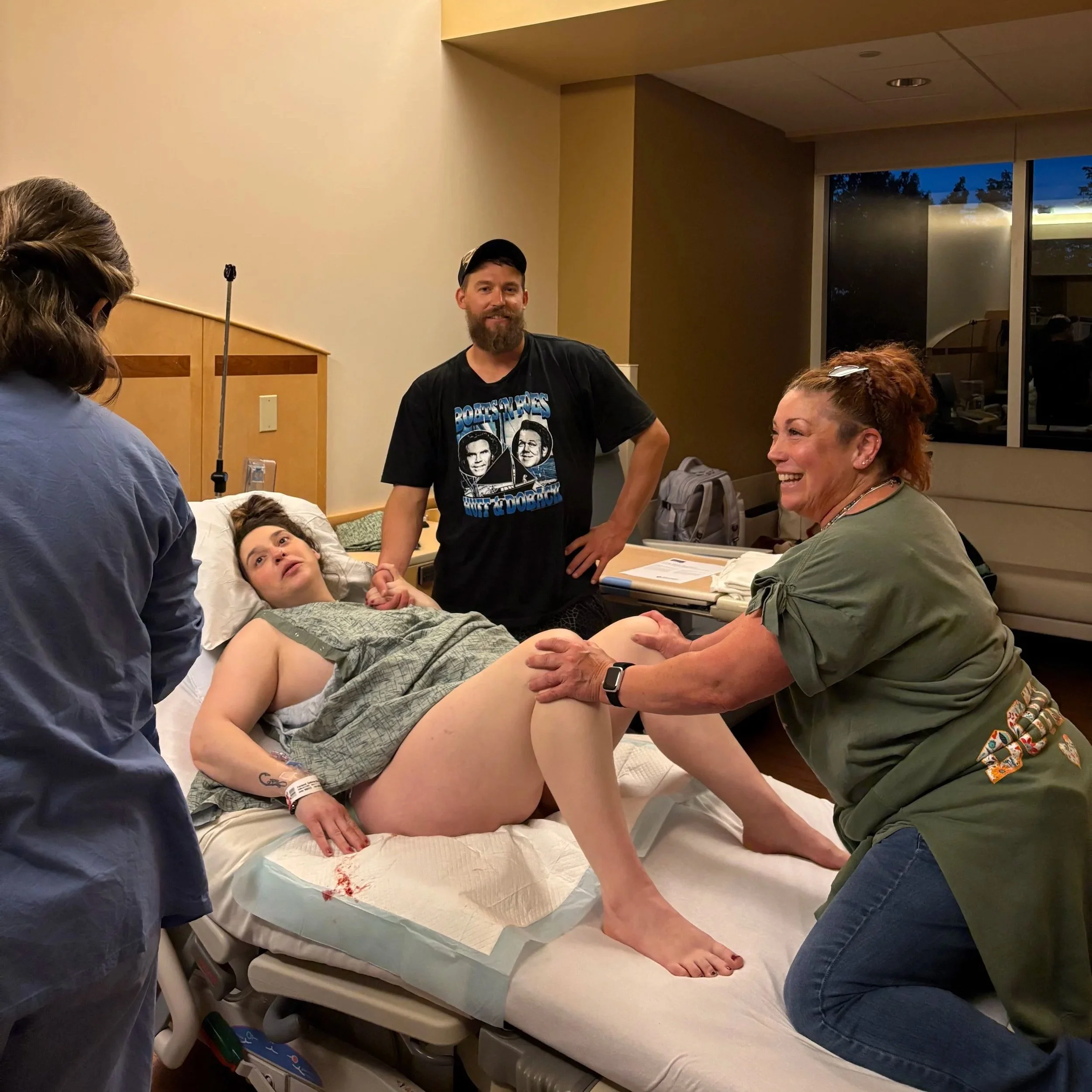 A woman laying in a hospital bed with a bloodstained sheet, surrounded by several people including a man standing beside her and a woman leaning over her, holding her hand and talking, in a hospital room.