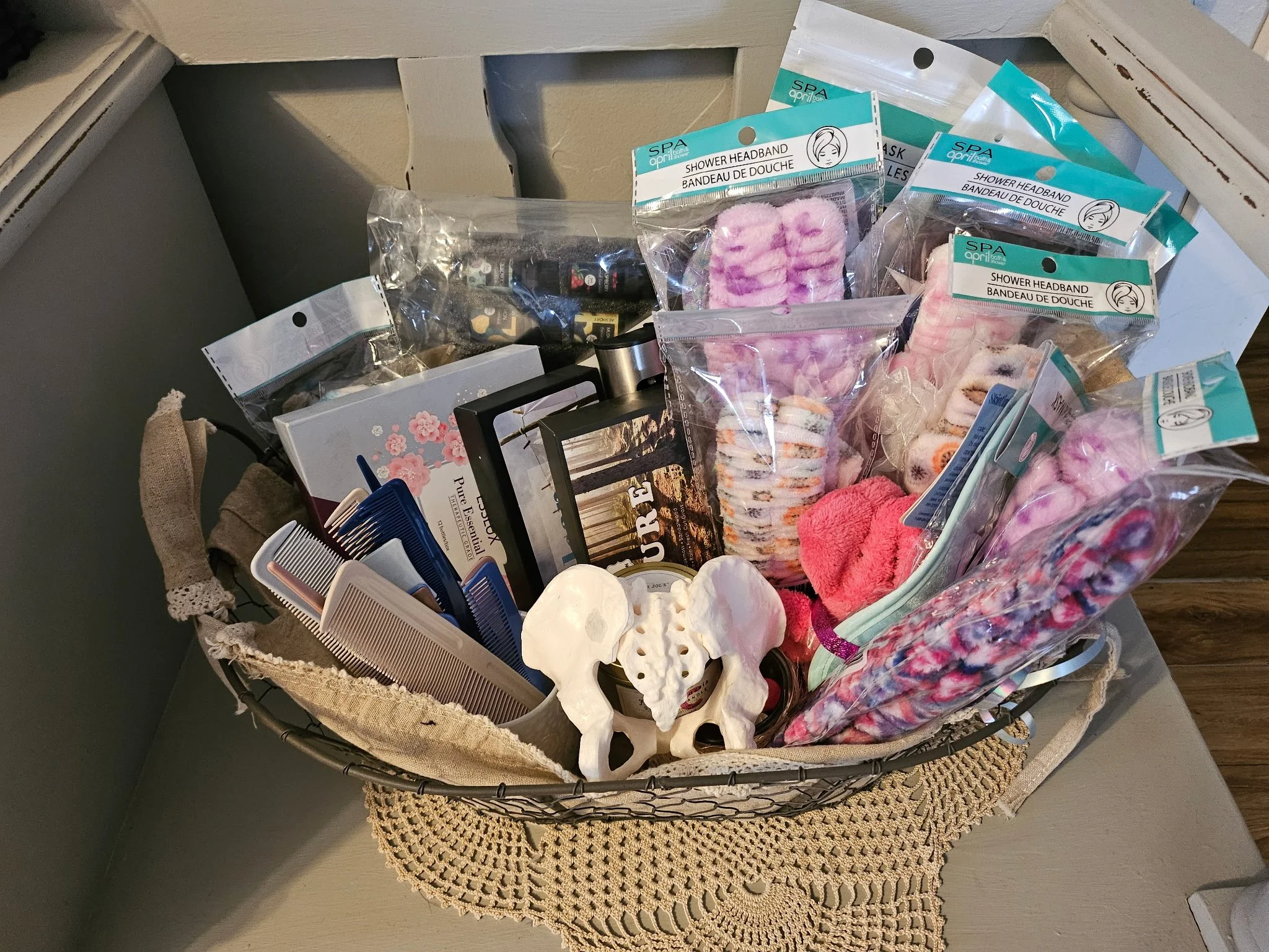 A basket contains various personal care and beauty items, including shower headbands, bath salts, a box of tissues, brushes, and other toiletries, placed on a table with a crocheted doily underneath.
