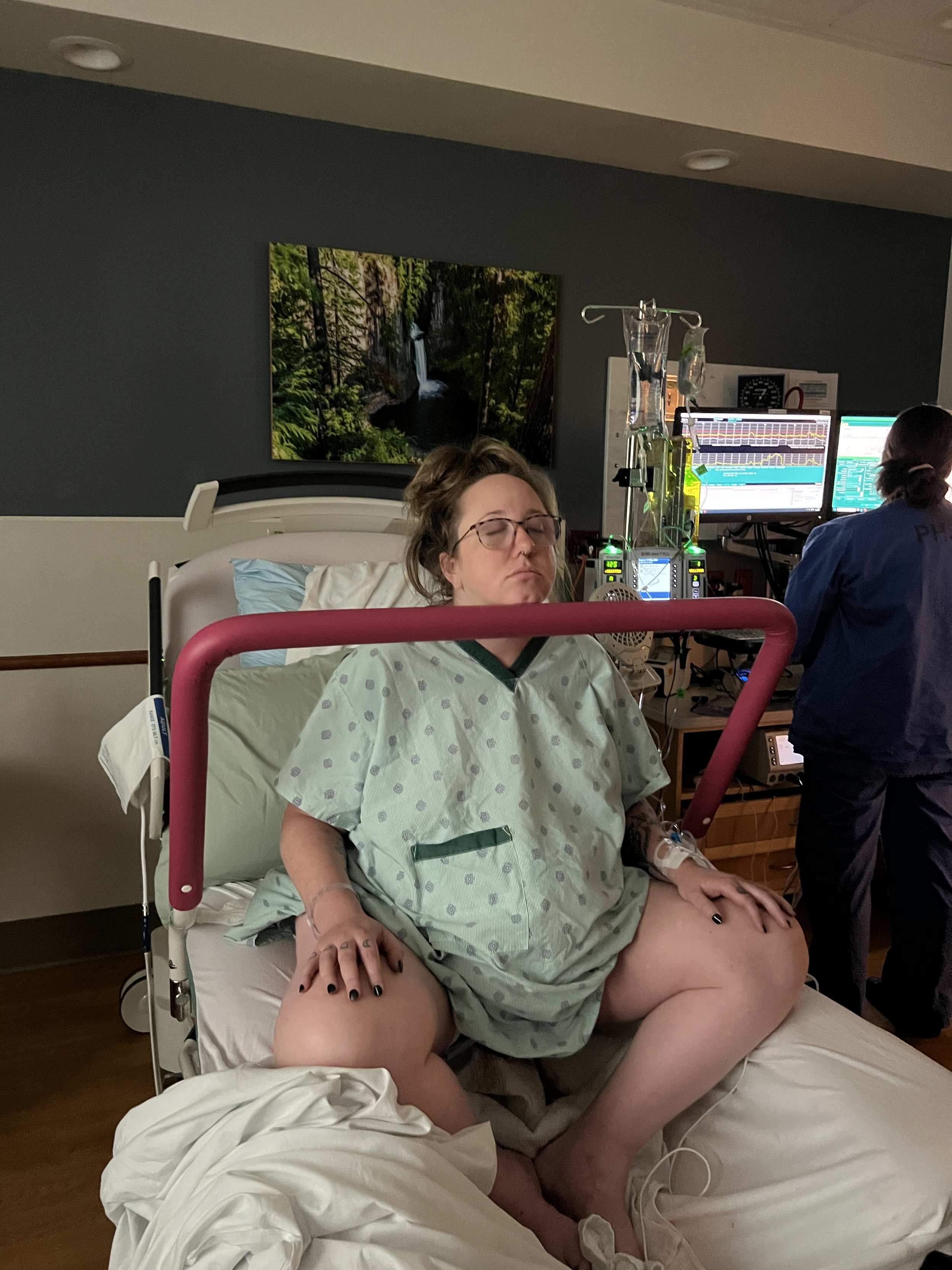 A woman lying in a hospital bed, with one hand on her thigh and her eyes closed, wearing a hospital gown. Medical equipment, monitors, and IV bags are visible in the background. A person, possibly a healthcare worker, stands next to her, wearing scrubs.