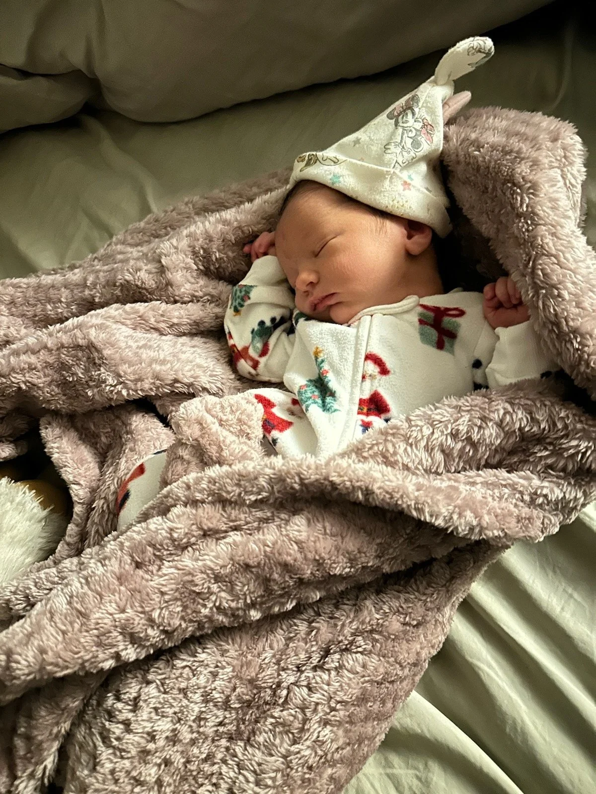 A sleeping baby wrapped in a soft, pink fuzzy blanket, wearing festive pajamas and a Christmas-themed hat with a pom-pom on top.