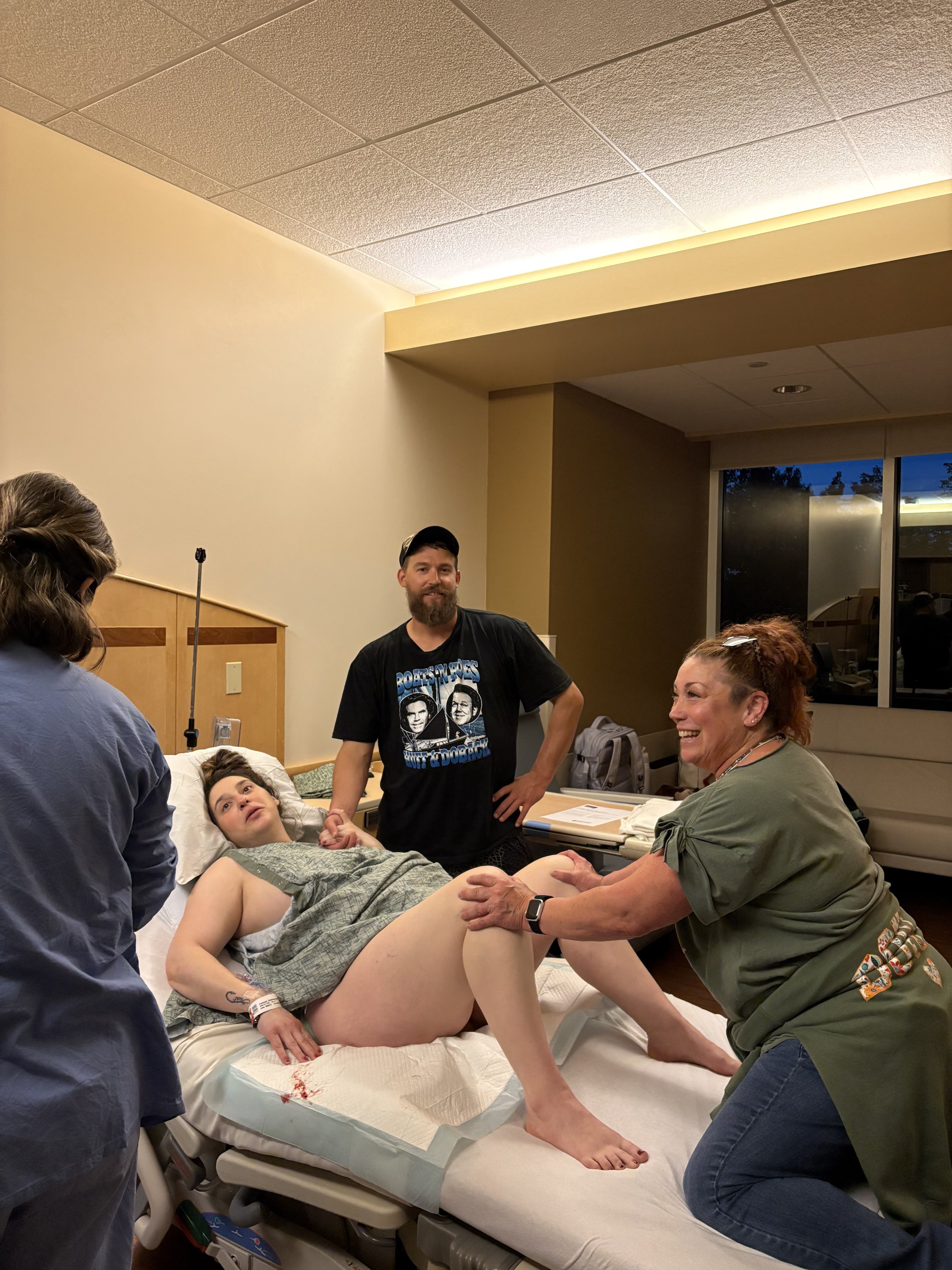 A woman lying on a hospital bed during labor, surrounded by a medical team including a doula. The birth doula is holding hands with someone off-camera, and a healthcare provider is supporting her leg.