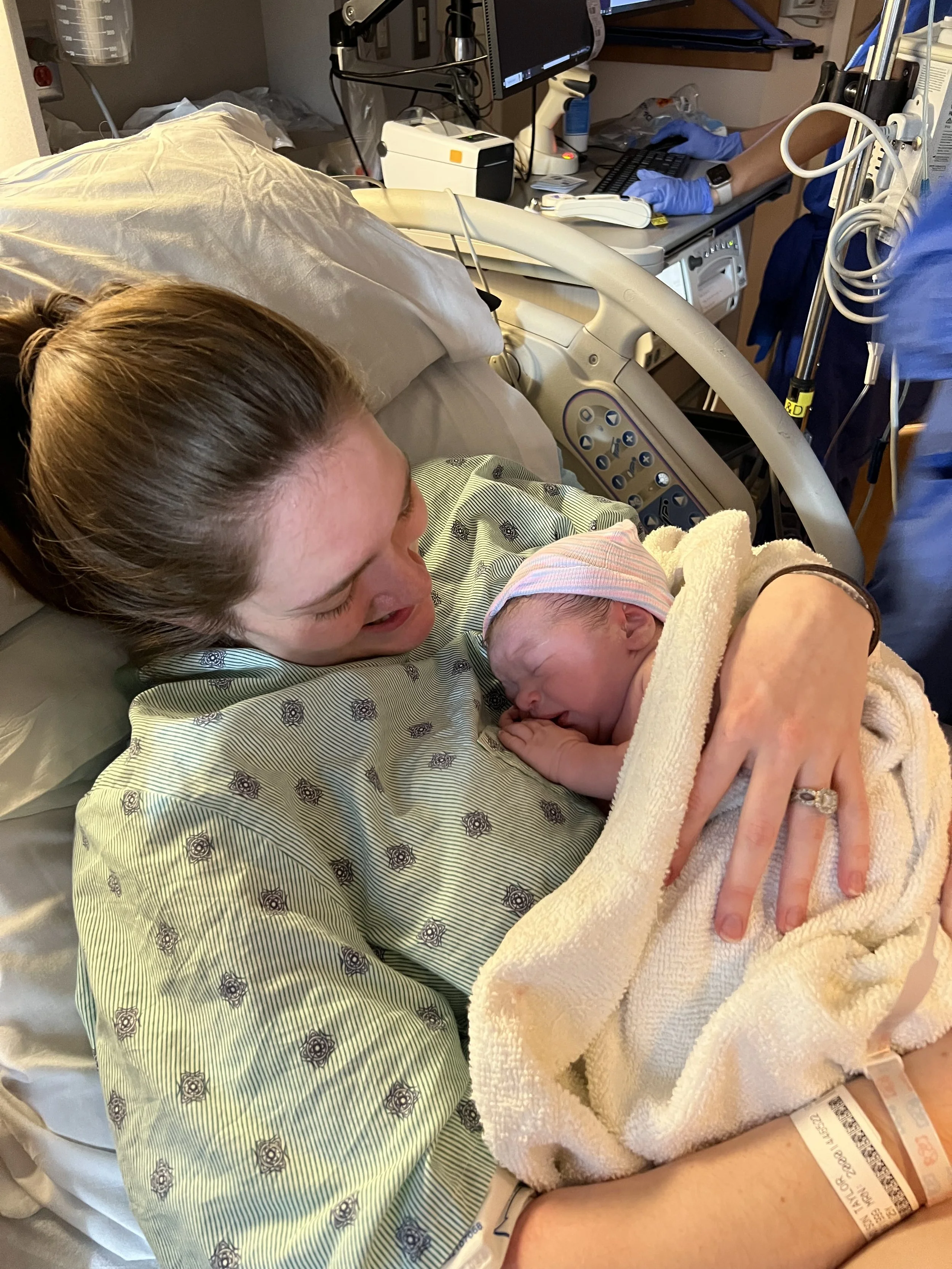 A woman in a hospital bed holding a newborn baby wrapped in a blanket. Medical equipment surrounds her, including IV bags, monitors, and healthcare staff in the background. Salem, Oregon Doula