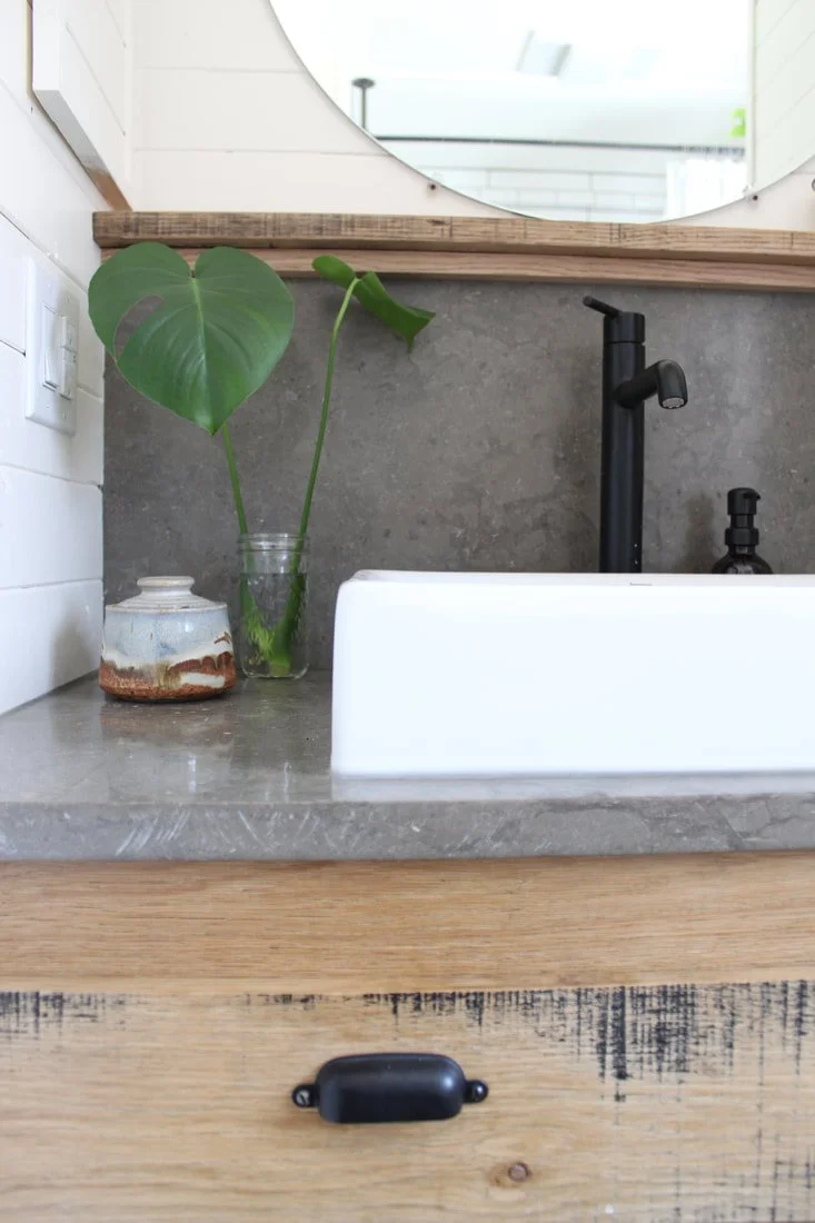 A bathroom countertop with a large leafy plant in a glass jar, a ceramic jar, a black faucet, a mirror, and a wooden cabinet with a black handle.