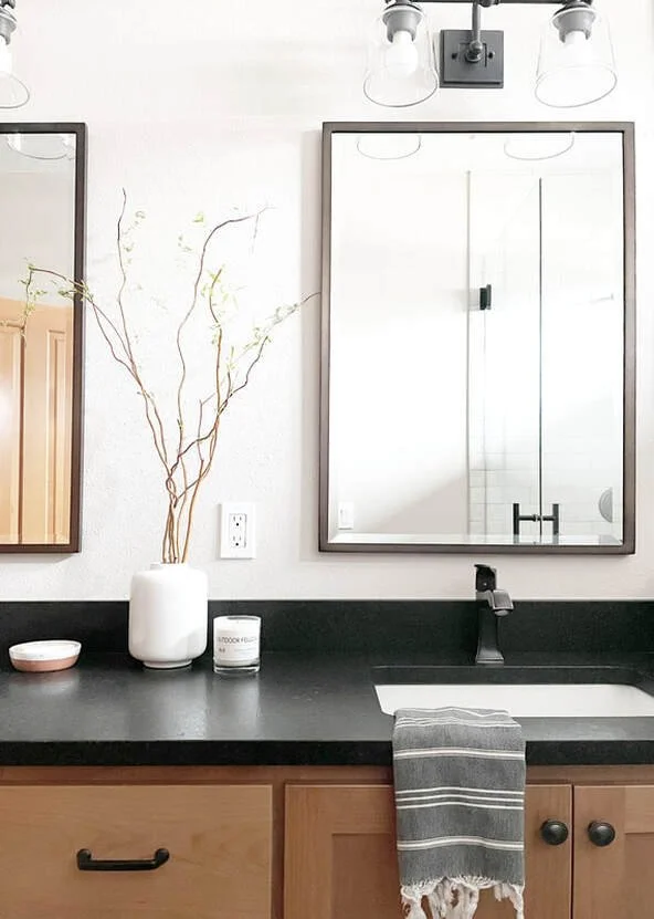 Bathroom vanity area with a black countertop, wooden cabinets, a white sink, a striped towel, a mirror, a vase with twigs, and a candle.