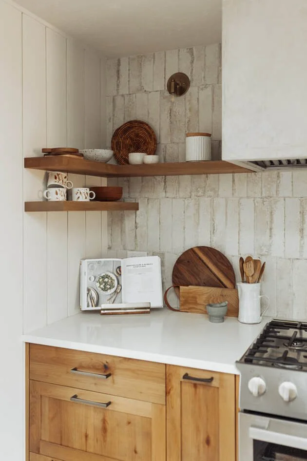 Open kitchen corner with wooden cabinets, white countertop, floating wood shelves with bowls and cups, cookbook, cutting board, utensils, and stove.