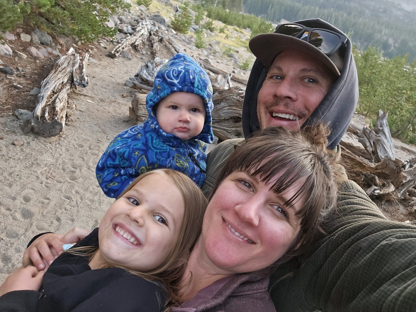 Family of four outdoors on rocky terrain, smiling at the camera. An adult man and woman are holding two young girls. The man wears a cap and hoodie, the woman has her hair tied up, and the girls are smiling happily. The background includes rocks, trees, and distant mountains.