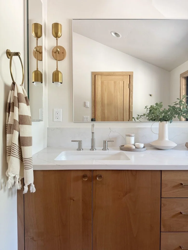 A bathroom vanity with a white countertop, wooden cabinets, and a large mirror. There are gold wall sconces, a striped towel hanging on a ring, and decorative items including a white vase with green foliage, candles, and stones.