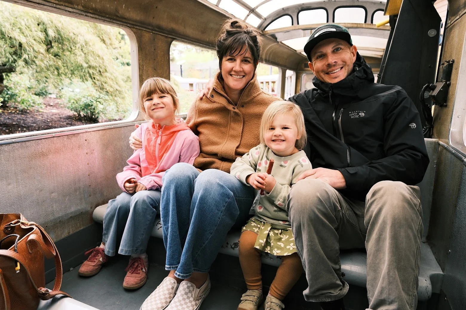 A family riding a train or tram, with two young girls and two adults, smiling and enjoying the ride in a vintage-style transportation vehicle. Outside greenery is visible through the windows.