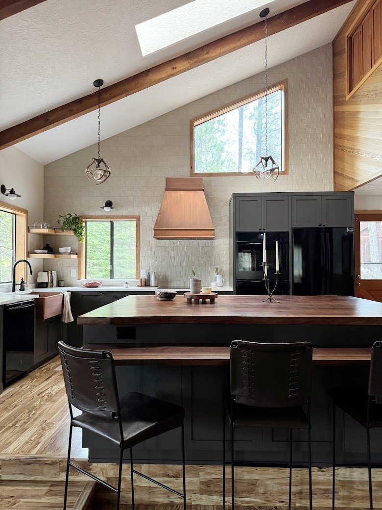 Modern kitchen with high vaulted ceiling, skylight, wooden beams, gray cabinets, black appliances, a large wooden island, pendant lights, and open shelving with plants and dishes.