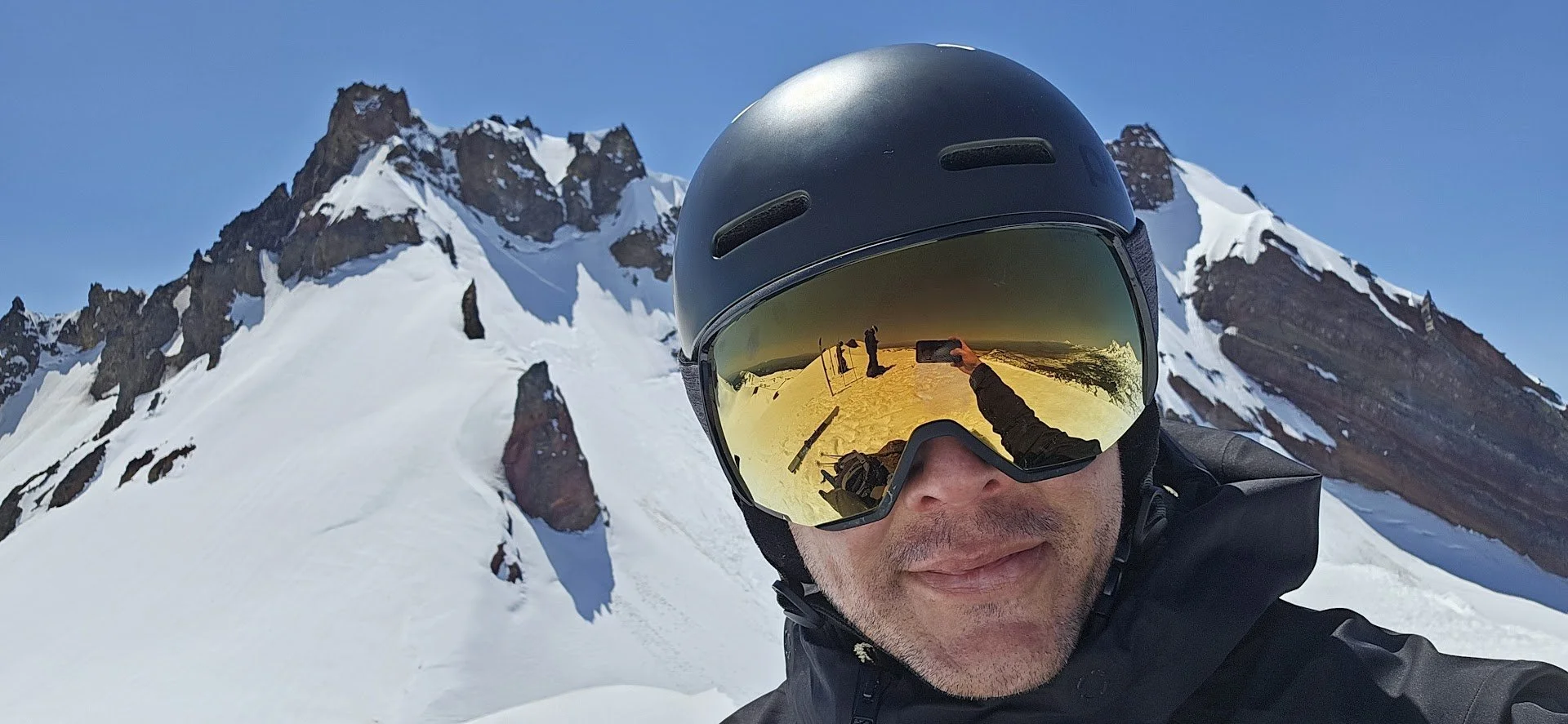 Person wearing a black helmet and ski goggles taking a selfie on snowy mountain slope with rocky peaks in the background.