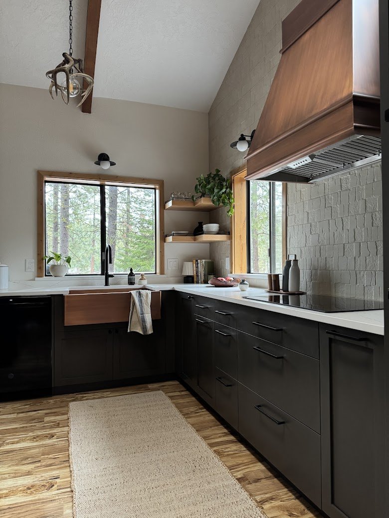Modern kitchen with black lower cabinets, white countertops, wooden window frames, open shelving with plants and kitchenware, a parking stove, gray backsplash, hardwood floor, and large windows showing trees outside.
