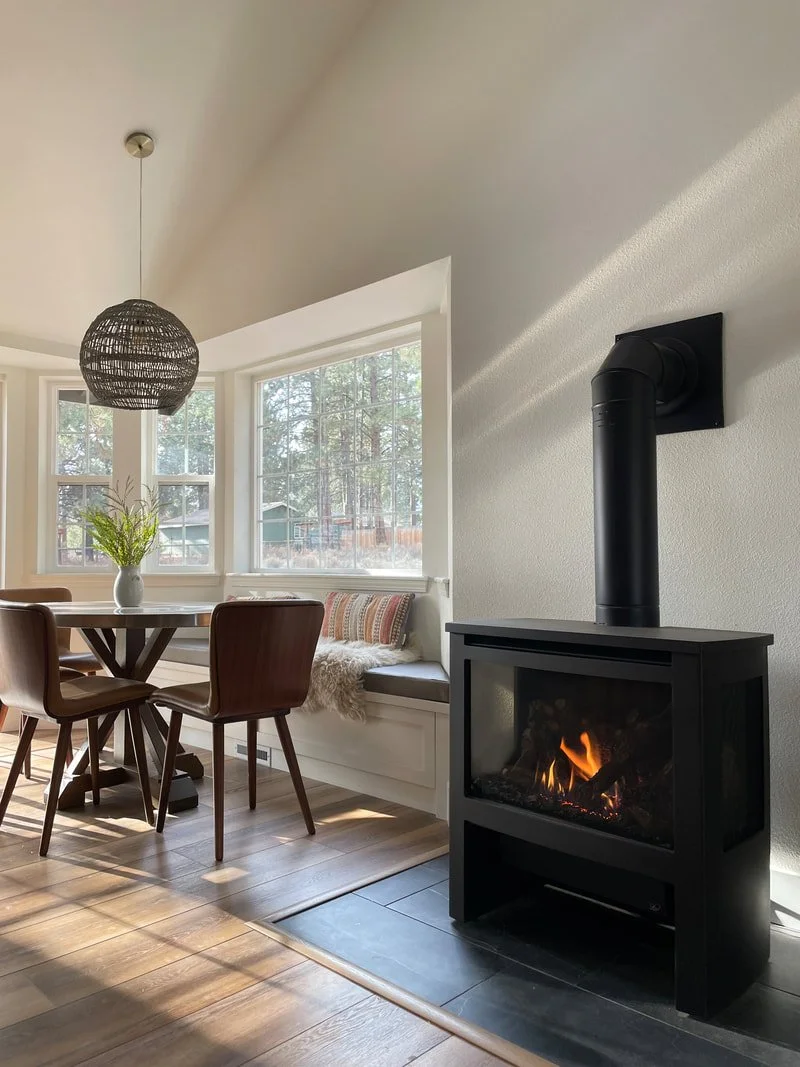 A cozy room with a dining area featuring a round wooden table, four wooden chairs, a vase with green leaves, and a bench with pillows and a blanket by the window. There is a black fireplace with a visible fire and a chimney on the wall to the right.