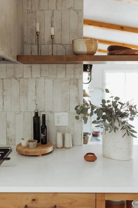 Kitchen countertop with a large white vase containing green and purple foliage, a small round terracotta dish, and a wooden tray holding bottles and small bowls. Behind the countertop is a beige tiled wall with electrical outlets and a nearby window 