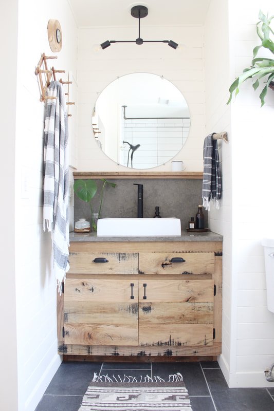 Bathroom with a rustic wooden vanity, round mirror, black faucet, and striped towels, with a plant on the left and a wall clock on the top left.