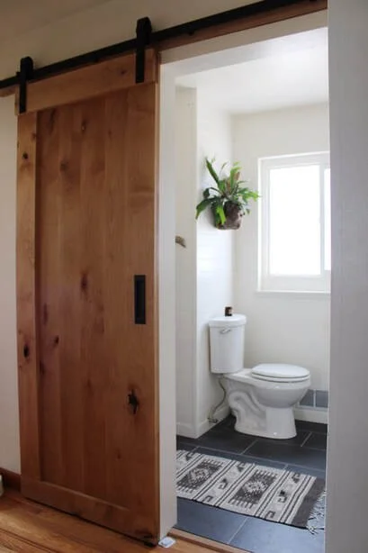 View of a small bathroom with a white toilet, a window, a hanging potted plant, and a sliding wooden door.
