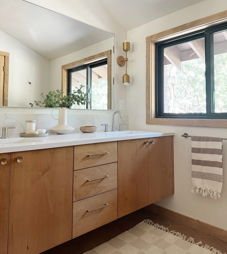 Bathroom vanity with a wooden cabinet, white countertop, and mirror, decorated with a white vase with greenery, a candle, and a small bowl, with a window and gold-colored wall sconce.