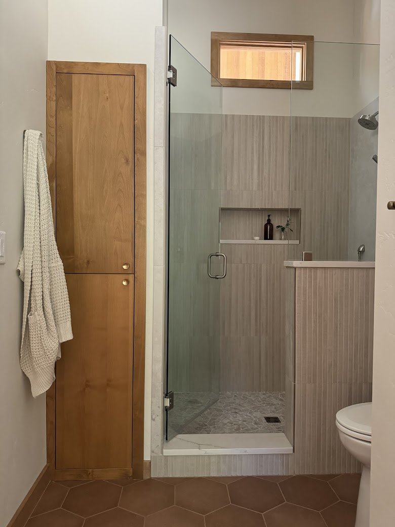 A bathroom with a wooden cabinet, a towel hanging on the wall, a glass shower with a small built-in shelf, and a toilet. There is a small window with a wooden frame above the shower.
