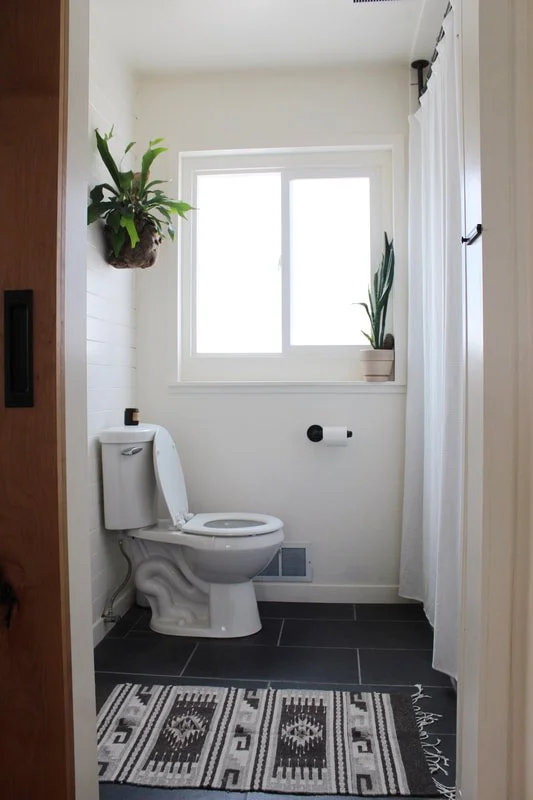 A small, bright bathroom with a toilet, a window with a white curtain, two potted plants on the windowsill and wall, a black and white patterned rug, and white walls and cabinetry.