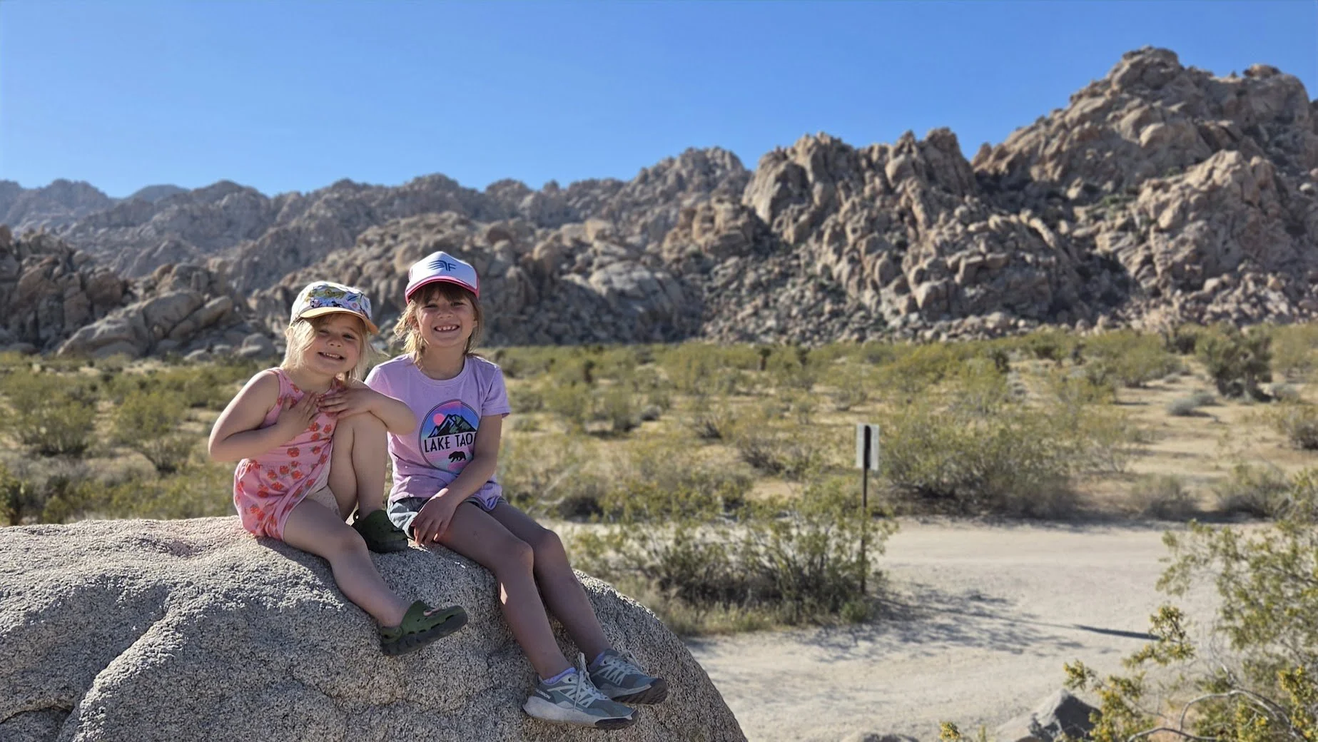 Two young girls sitting on a large rock in a desert landscape with mountains in the background, wearing casual summer clothes and hats, smiling at the camera.