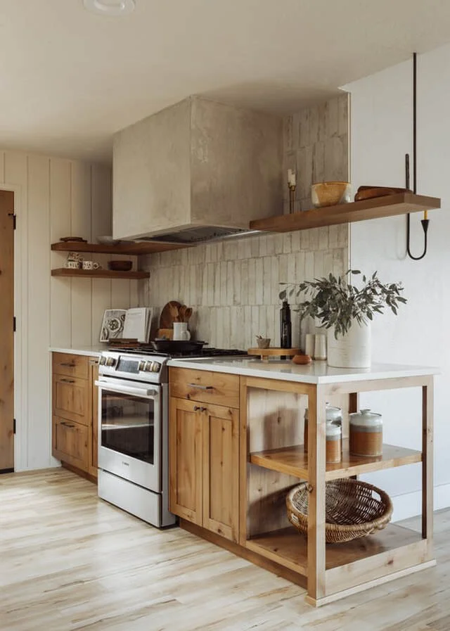 A cozy kitchen with wooden cabinets, open wooden shelves, a white countertop, and a small open shelving unit with jars and a basket. The background features a rustic, partial brick wall behind the stove and a large white pot with a green plant on the